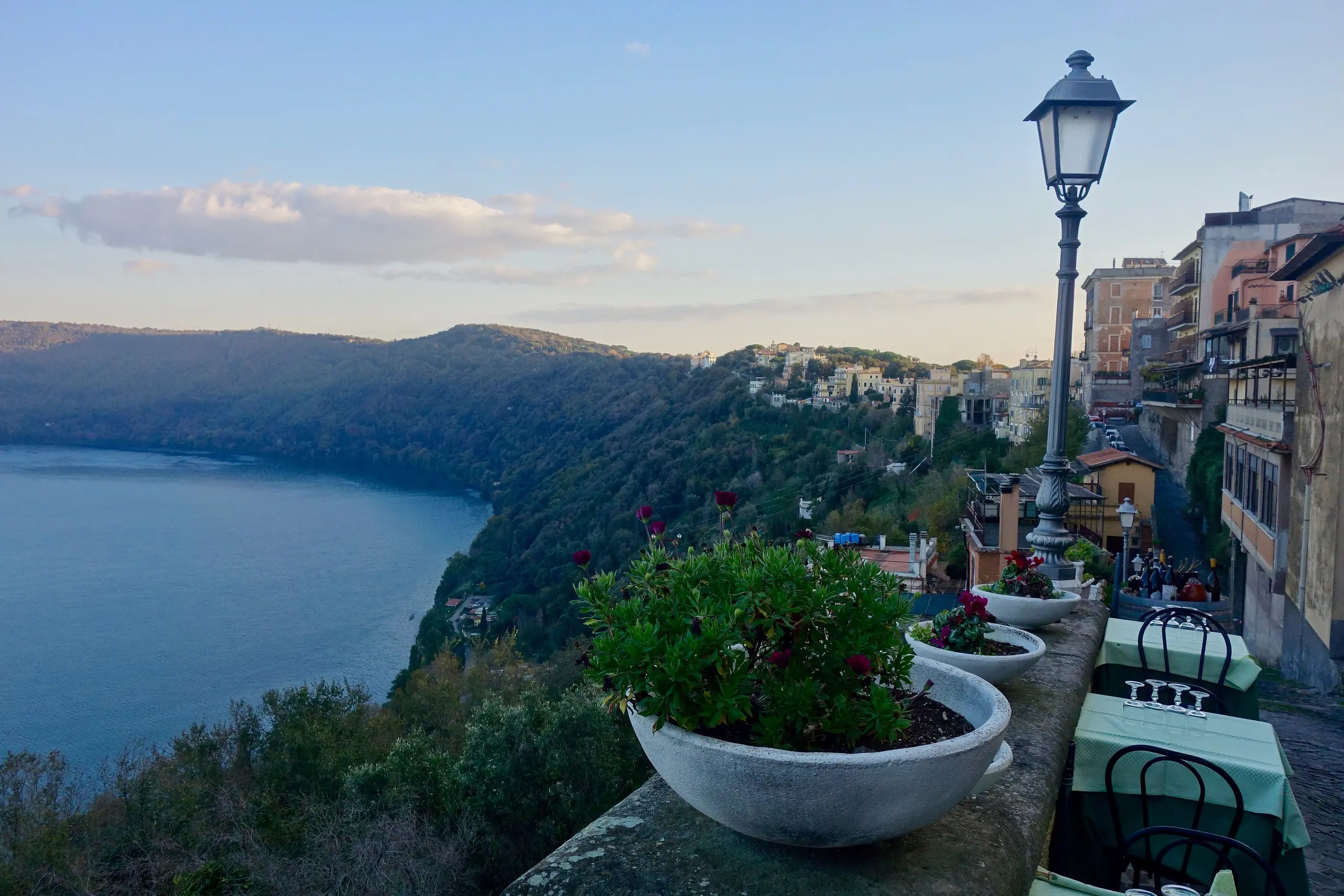A café terrace in Castel Gandolfo overlooking the blue crater of Lake Albano at dusk, with a street lamp and flowering pots.