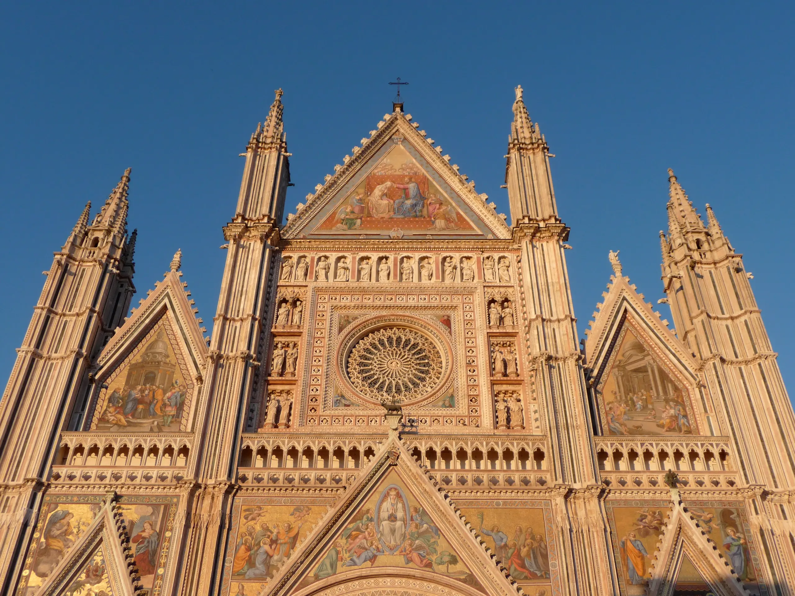 The gabled facade of Orvieto Cathedral glowing gold in late light, its mosaics and spires against a deep blue sky.