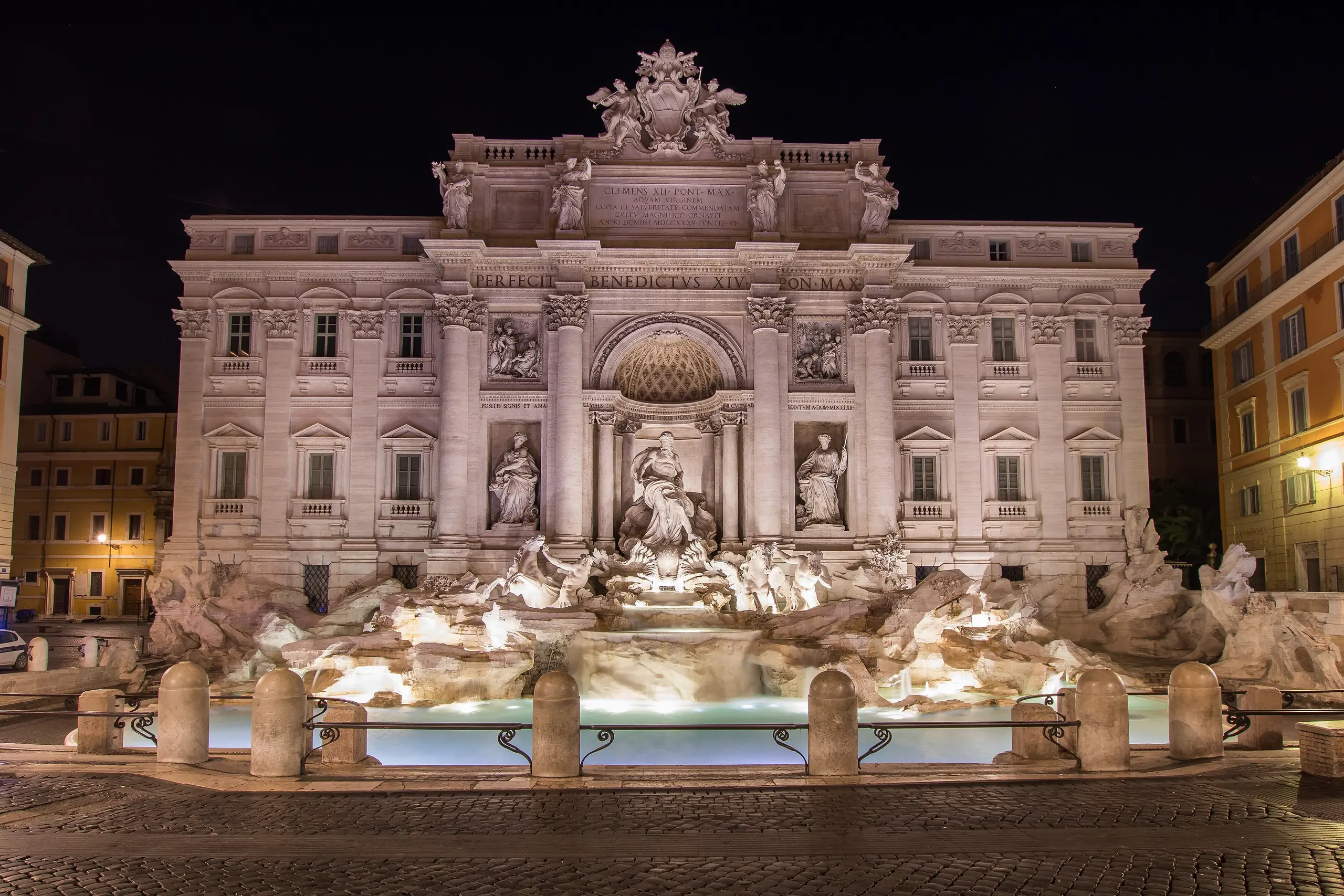 The Trevi Fountain lit at night across an empty, cobbled Piazza di Trevi, its facade glowing against a black sky.